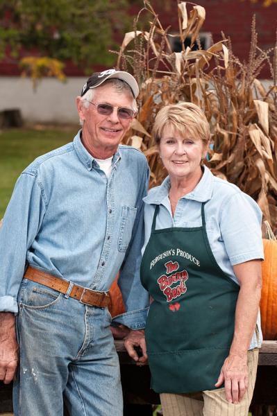 A man and a woman standing in front of corn stalks and pumpkins 